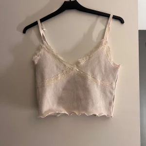 Beige ribbad linnetopp från Divided - Säljer en söt beige ribbad linnetopp från Divided med spetsdetaljer. Toppen har smala axelband och en v-ringad design. Perfekt för varma dagar eller som ett lager under en tröja.