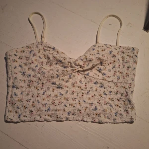 Blommig linnetopp från Even&Odd - Söt blommig linnetopp från Even&Odd med smala axelband och en knytdetalj framtill. Toppen har ett litet blommönster i flera färger på en vit bakgrund. Perfekt för varma sommardagar!