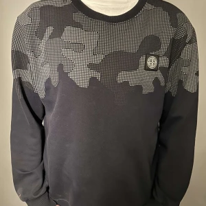 Svart camouflagemönstrad tröja från Stone Island - Säljer en svart tröja från Stone Island med ett coolt camouflagemönster i grått. Tröjan har långa ärmar och en rund halsringning. Perfekt för en stilren look.