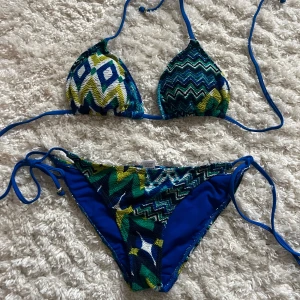 Mönstrad missoni bikini i blå och gröna toner - Snygg bikini med zigzag-mönster i blå, gröna och vita nyanser. Toppen har justerbara knytband och trekantsformade kupor. Bikinitrosan har knytband i sidorna för perfekt passform. MISSONI LIKNANDE, köpt i Positano Italien för 55 euro. 