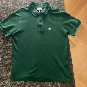 Lacoste piké - Säljer en grön pikétröja från lacoste. Pikén har korta ärmar och en knappslå med tre knappar. Perfekt för en stilren och avslappnad look.