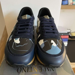 Valentino Rockrunner  - Snygga Valentino Rockrunner sneakers med ett coolt camouflagemönster i blå, svart och beige. Skorna har snörning och en unik design med detaljer i skinn och syntet. Perfekta för att ge din outfit en trendig touch. priset är ej fast i ett sten