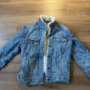 Jeansjacka med teddyfoder från Levi's -  en jeansjacka från Levi's med mjukt vitt teddyfoder. Jackan har en snygg blå tvätt och knappar framtill. Perfekt för kyligare dagar och stilrena design. passar både s ny pris 900