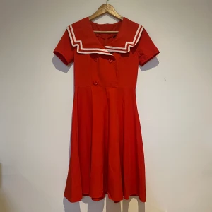 Röd klänning med sjömanskrage - Obs!! En knapp saknas men är ej något märkbart. Charmig röd klänning med en unik sjömanskrage i vitt. Klänningen har korta ärmar och knappar framtill som ger en retro känsla. Perfekt för en somrig look med sin livfulla färg och klassiska design.