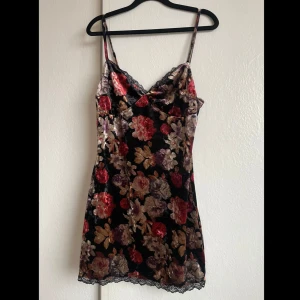 Blommig klänning från Narrated - Snygg blommig klänning från Narrated i svart med färgglada blommor. Klänningen har smala axelband och spetsdetaljer vid halsringningen. Tajtare passform 