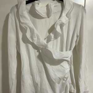 Vit omlotttopp med volangdetaljer - Säljer en stilren vit omlottopp med volangdetaljer. Toppen har långa ärmar och en elegant omlottdesign med utsvängda ärmslut. Den är i storlek small men skulle själv säga att den sitter mer som medium.