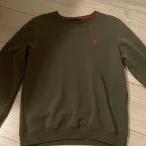 Olivgrön tröja från Polo Ralph Lauren - Säljer en stilren olivgrön tröja från Polo Ralph Lauren med det klassiska röda logotypbroderiet på bröstet. Tröjan har långa ärmar och en rund halsringning med ribbade kanter.