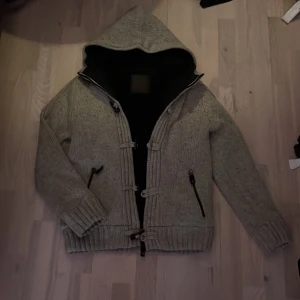 Grå stickad cardigan - Säljer en grå stickad cardigan med huva och dragkedja. Jackan har ribbade muddar och två fickor med dragkedja framtill. Insidan är fodrad med ett mjukt material i päls för extra värme. Perfekt för kyliga dagar! True to size!
