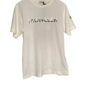 Moncler tshirt storlek S  - Moncler t-shirt i storlek S. Klassisk och stilren design i vit färg, tillverkad av högkvalitativa material som kombinerar komfort och lyx. T-shirten är i nyskick och kommer med original prislapp kvar. Perfekt för en avslappnad men exklusiv look. En tidlös basplaggsinvestering från det ikoniska Moncler.
