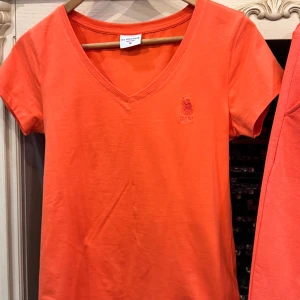 Orange mjukisbyxor från U.S. Polo Assn. - Snygga orange mjukisbyxor från U.S. Polo Assn. med broderad logga 'USPA' i flera färger på benet. Byxorna har en bekväm passform med snörning i midjan för justerbarhet. Perfekta för en avslappnad stil.
