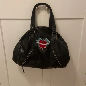 Svart väska med broderat hjärta och små detaljer  - Snygg svart handväska i skinn med ett coolt broderat hjärta med vingar på framsidan. Väskan har två dragkedjor framtill och en mycket rymlig insida med extra fack. Nypris ligger runt 400-700kr. Pris kan diskuteras!💗