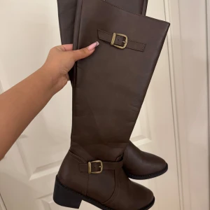Bruna stövlar med spänne - Snygga bruna stövlar i skinn med dekorativa spännen. Perfekta för höst och vinter, dessa stövlar har en klassisk design med låg klack och rund tå.