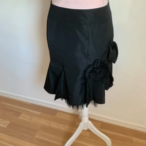 Svart kjol med volangdetaljer - Elegant svart kjol med unika volangdetaljer på sidan. Kjolen har en klassisk passform och är perfekt för att ge en stilfull touch till din outfit. Den är knälång och har en subtil glans i materialet.