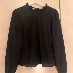 Svart chiffongblus med volang - Säljer en stilren svart chiffongblus från H&M Divided. Blusen har långa ärmar och en fin volangdetalj vid halsen och ärmarnas slut. Den har knytning i ryggen. Perfekt för att klä upp en vardagsoutfit eller för en kväll ute. Säljer i storlek 38 men är ganska liten i storlek.