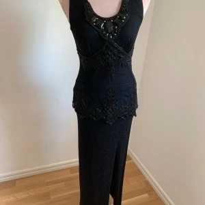 Svart långklänning med pärldetaljer - Elegant svart långklänning med vackra pärldetaljer och spetsmönster. Klänningen är ärmlös och har en figursydd passform som framhäver midjan. Perfekt för en glamorös kväll!