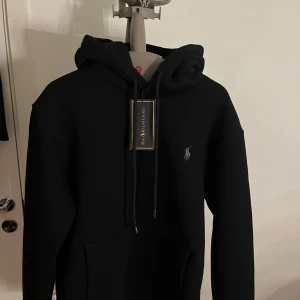 Svart hoodie från Ralph Lauren - Säljer en stilren svart hoodie från Ralph Lauren med det klassiska logotypbroderiet på bröstet. Hoodien har en justerbar dragsko i huvan och en praktisk magficka. Perfekt för en avslappnad look.