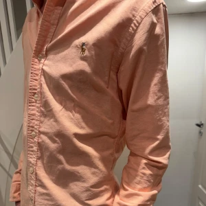 Persikofärgad skjorta från Ralph Lauren - Snygg persikofärgad skjorta från Ralph Lauren med klassisk krage och knappar framtill. Skjortan har en broderad logotyp på bröstet och långa ärmar. Perfekt för en stilren look.