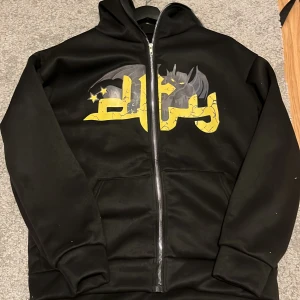 Svart hoodie med tryck - Säljer en svart hoodie med dragkedja och ett coolt tryck av en fladdermus och stjärnor i grått och gult på framsidan. Hoodien har långa ärmar och en bekväm passform. Perfekt för en avslappnad stil.