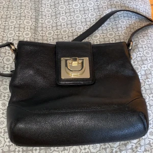 Svart axelväska från DKNY - Snygg svart axelväska från DKNY i skinn med en elegant gulddetalj på framsidan. Väskan har en justerbar axelrem och ett rymligt fack med dragkedja inuti. Perfekt för att bära dina dagliga nödvändigheter med stil.