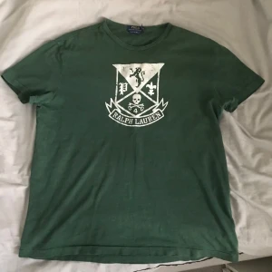 Grön t-shirt från Ralph Lauren - Säljer en grön t-shirt från Ralph Lauren med ett stort tryck på framsidan. T-shirten har en klassisk passform och är kortärmad. Perfekt för en avslappnad stil.