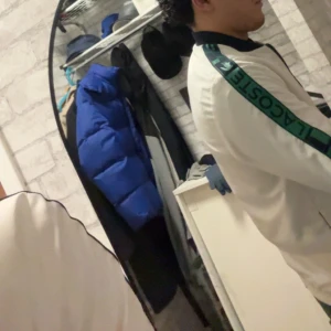 Vit windbraker från Lacoste - Säljer en snygg vit jacka från Lacoste med gröna detaljer och logotyp på ärmen. Jackan har en klassisk design med dragkedja och är perfekt för en sportig look. Den är långärmad och har en bekväm passform.