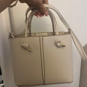 Beige handväska med justerbar axelrem - Snygg beige handväska med justerbar axelrem och gulddetaljer. Väskan har en stilren design med två handtag och en praktisk dragkedja upptill. Perfekt för att bära dina dagliga nödvändigheter med stil.