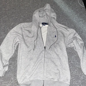 Grå hoodie från Polo Ralph Lauren - Säljer en stilren grå hoodie från Polo Ralph Lauren med dragkedja och den klassiska loggan broderad på bröstet. Tröjan har långa ärmar och en praktisk huva med dragsko. Perfekt för en avslappnad stil.helt ny storleken S/M