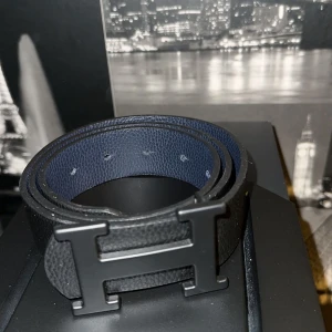 Svart bälte med H-spänne - Snyggt svart bälte med ett stort H-format spänne. Perfekt för att ge en stilren touch till din outfit. Bältet har en slät yta och flera hål för justering.