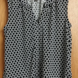 Svartvit ärmlös topp med geometriskt mönster - Snygg svartvit ärmlös topp med ett geometriskt mönster. Tillverkad i polyester och elastan för en bekväm passform. Perfekt för en stilren look.