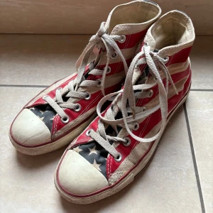 Converse med amerikanskt flaggmönster - Snygga Conversesneakers med ett amerikanskt flaggmönster i rött, vitt och blått. Skorna har klassisk snörning och den ikoniska Chuck Taylor-loggan på sidan. ————————————— Välanvända med slitage i hälen och sulan.
