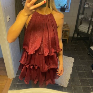 Vinröd klänning med volanger - Klänningen är helt oanvänd! Säljer en vinröd klänning med flera lager av volanger. Klänningen är ärmlös och har en luftig passform, perfekt för festliga tillfällen. 