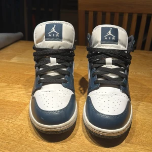 Nike Air Jordan 1 Mid Armory Navy - Snygga Nike Air Jordans i en stilren kombination av blått och vitt. Skorna har klassisk snörning och den ikoniska Air Jordan-loggan på sidan. Perfekta för dig som vill ha en sportig och trendig look. Storlek 40. Inköpta för 2 år sedan. Använt skorna ett flertal gånger. Lite Creasade på bild 1. Ett pytte litet märke på vänstra skon vid bild 1 vid sulan.
