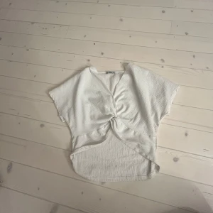 Vit ribbad topp - Säljer en vit ribbad topp med korta ärmar och en knutdetalj framtill. Perfekt för en avslappnad stil. Toppen har en lös passform och är tillverkad i ett mjukt material.