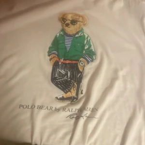 Vit t-shirt med Polo Bear från Ralph Lauren - Vit t-shirt från Ralph Lauren med ett tryck av den ikoniska Polo Bear i en grön jacka och randig tröja. Perfekt för en avslappnad stil med en touch av lyx. Ny pris mellan 800 - 1200kr