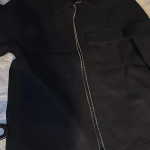 Svart jacka med dragkedja - Säljer en stilren svart jacka med dragkedja framtill. Jackan har en klassisk krage och en praktisk bröstficka. Perfekt för en minimalistisk look.