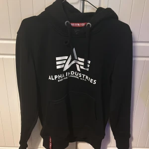 Svart hoodie från Alpha Industries - Snygg svart hoodie från Alpha Industries med deras ikoniska logga tryckt på framsidan. Tröjan har en klassisk känguruficka, riktigt skön tröja, har använt den några få gånger inga märken så som färg eller smuts.