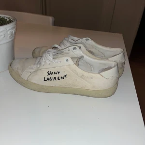 Vita sneakers från Saint Laurent - Säljer ett par vita sneakers från Saint Laurent med svart text på sidan. Skorna har en klassisk design med snörning och en bekväm sula. Perfekta för en stilren look.