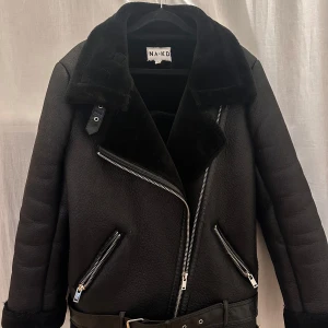 Svart jacka från NA-KD - Snygg svart jacka från NA-KD med dragkedja och bälte. Jackan har en mjuk krage och är perfekt för kyligare dagar. Den har en cool bikerstil med två dragkedjefickor framtill.