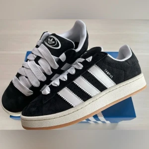 Svarta Adidas Campus sneakers - Snygga svarta Adidas Campus sneakers med vita detaljer och klassiska tre ränder på sidan. Skorna har en vit sula och breda vita snören. Perfekta för en stilren look.