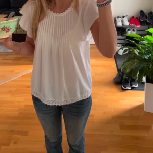 Vit blus med spetsdetaljer - Säljer en söt vit blus med spetsdetaljer vid hals och nederkant. Blusen har korta ärmar med volang och fina veck framtill. Perfekt för en somrig look! Storlek 42 men passar mig med S!💕