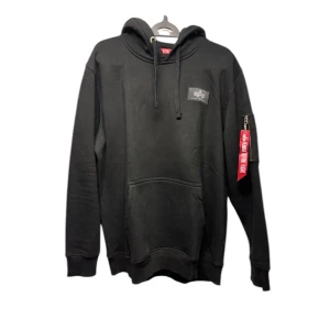 Svart hoodie från Alpha Industries - Snygg svart hoodie från Alpha Industries med stor logga på ryggen och en praktisk ficka med dragkedja på ärmen. Den har en klassisk känguruficka framtill och justerbar huva med snören. Perfekt för en casual look. Har använt rätt många gånger.