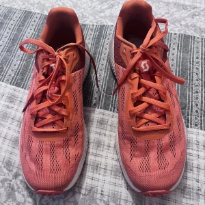 Orange träningsskor från Scott - Säljer ett par snygga orange träningsskor från Scott. Skorna har en ventilerande meshdesign och snörning för optimal passform. Perfekta för löpning eller gymmet med en slitstark sula för bra grepp. Skorna har en modern look med vita detaljer på sulan.