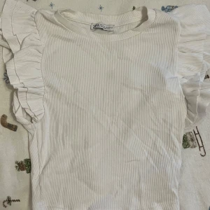 Vit topp med volangärmar från Zara - Säljer en söt vit topp från Zara med volangdetaljer på ärmarna. Säljer då den inte kommer till användning. Den är använd några få gånger 