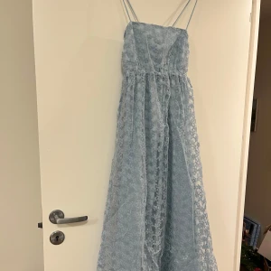 Ljusblå långklänning från NA-KD - Säljer en vacker ljusblå långklänning från NA-KD med blommigt mönster och smala axelband. Klänningen har en elegant snörning i ryggen och är perfekt för speciella tillfällen. Den är i ett skirt material som ger en romantisk känsla. Passar perfekt för vår och sommar.
