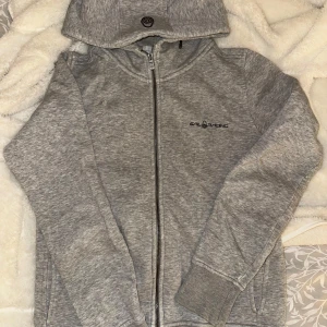 Grå hoodie från Sail Racing - Säljer en grå hoodie från Sail Racing med dragkedja och huva. Tröjan har långa ärmar och ribbade muddar. Den har ett hål vid vänstra ”brösten” men syns inte så mycket och någon liten defekt vid armen