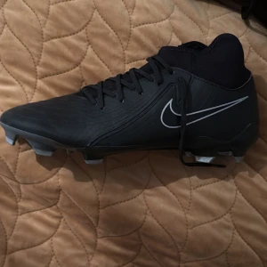 Svarta fotbollsskor från Nike - Snygga svarta fotbollsskor från Nike med snörning och en stilren design. Skorna har en slitstark sula med dobbar för bra grepp på planen. Perfekta för fotbollsspelare som vill ha både stil och funktion.