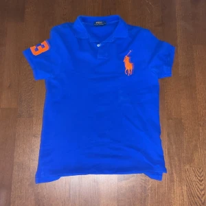 Blå pikétröja från Ralph Lauren - Snygg blå pikétröja från Ralph Lauren med orange logga och nummerdetalj på ärmen. Tröjan är slim fit och kortärmad, perfekt för en stilren look. Klassisk krage och knappar framtill.