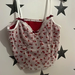 Vit och röd linne med körsbärsmönster - Charmig vit linne med rött körsbärsmönster och spetsdetaljer vid halsen. Toppen har smala axelband och en söt rosett framtill. Perfekt för en somrig look! Finns 3 st av denna.