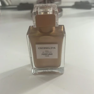 Cocosiliya Eau de Parfum - Elegant parfymflaska från Cocosiliya med en stilren design. Flaskan är guldig, med en fyrkantig, transparent kork. Etiketten är vit med svart text som ger en lyxig känsla. Perfekt för den som söker en sofistikerad doft. Parfymen kommer inte till användning då jag har många andra parfymer.