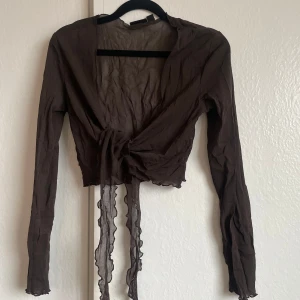 Brun transparent topp från Lindex - Säljer en brun transparent topp från Lindex med långa ärmar och knytband framtill. Toppen har volangdetaljer längs kanterna och är perfekt för en trendig look. Litet hål uppe vid lappen